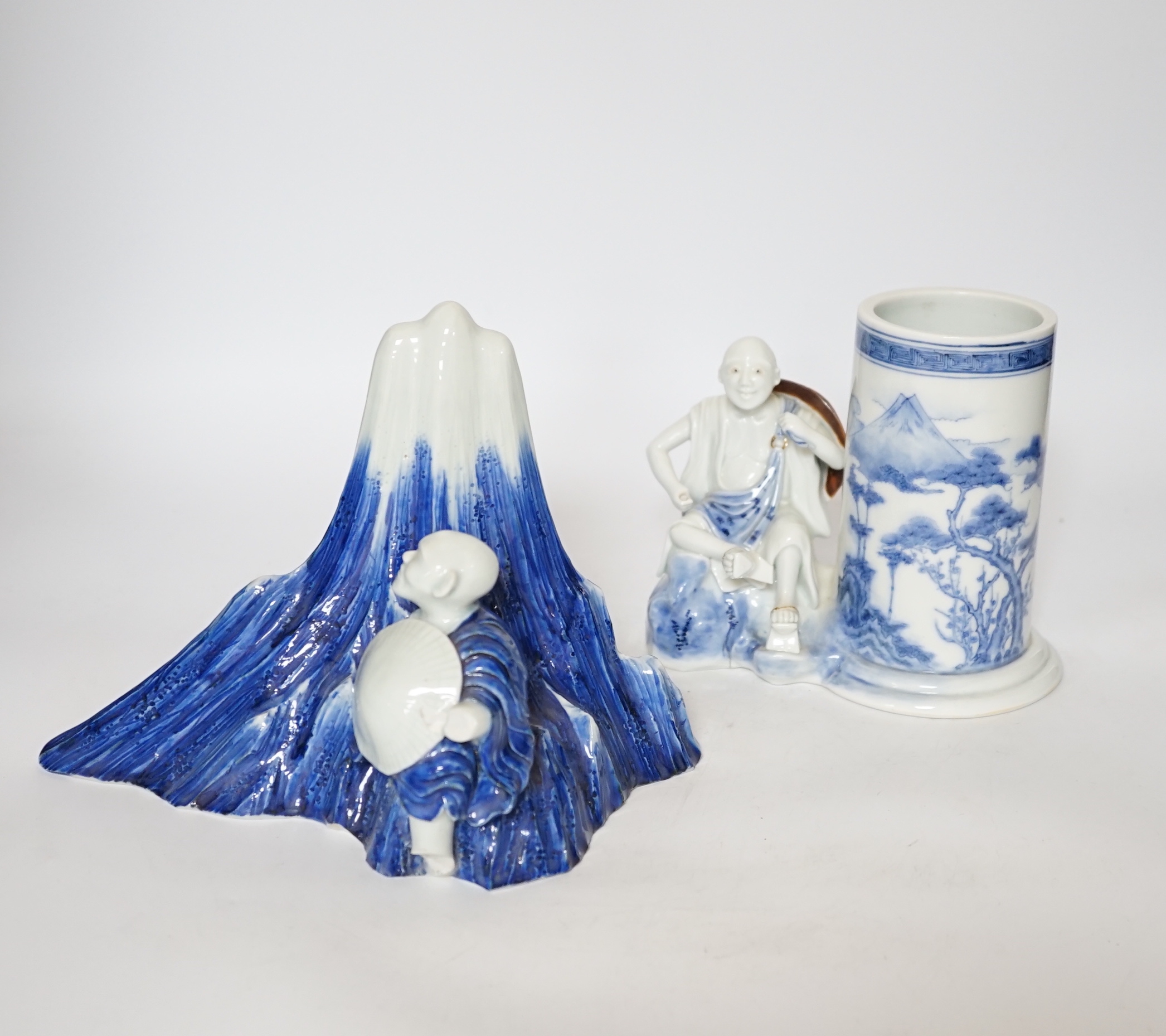 An early 19th century Japanese Hirado blue and white model of a monk seated by Mount Fuji, and a similar brush pot modelled with a fisherman seated by the cylindrical vessel, largest 17cm high (2)